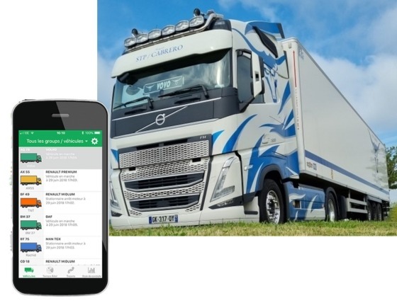 Un grand semi-remorque Volvo bleu et blanc est garé sur l'herbe sous un ciel bleu. Au premier plan, un smartphone affiche une application de suivi de véhicules électriques pour la gestion de flotte.