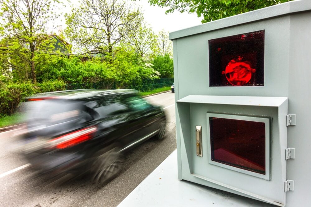 A black car speeds past a roadside traffic camera with a red flash, indicating the vehicle has been photographed for speeding, highlighting the importance of sicheres Fahrverhalten.
