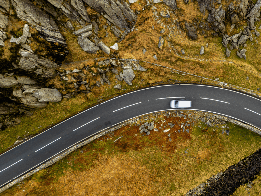 Aerial view of a white car driving on a winding mountain road bordered by rocky terrain and patches of grass.