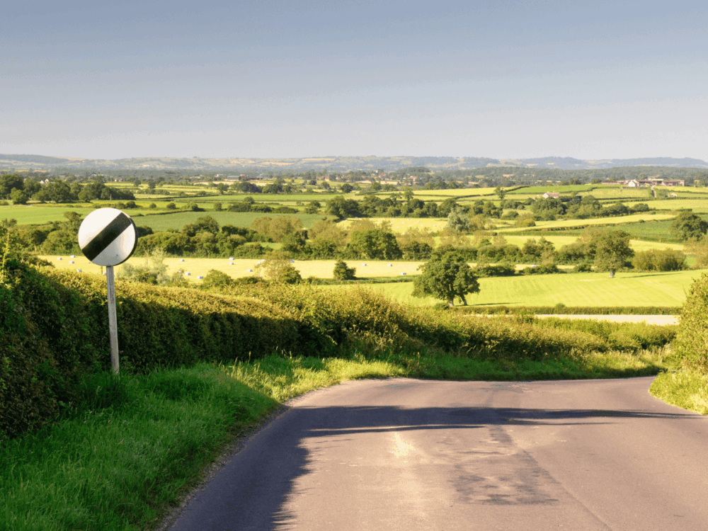 A narrow country road curves through a green rural landscape under a clear sky, with a UK national speed limit sign on the left-hand side.