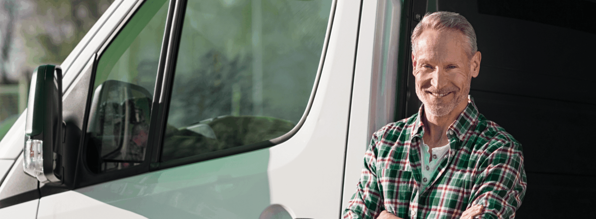 A man in a plaid shirt stands with arms crossed and smiles beside a white van on a sunny day.