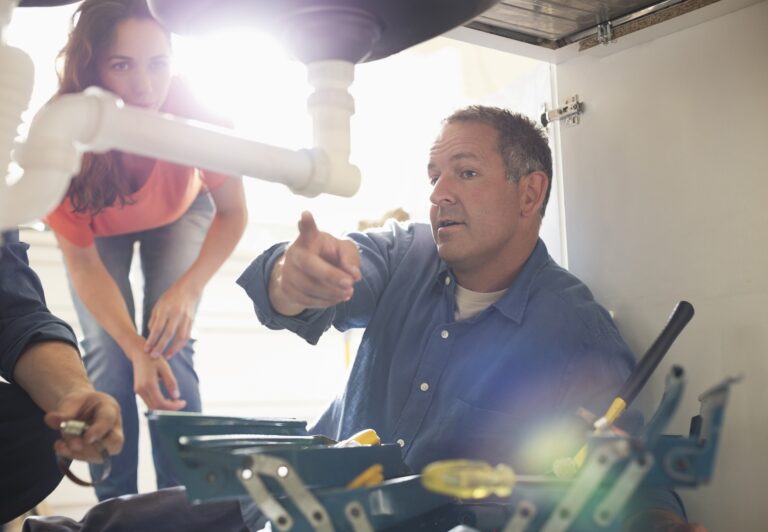 A man working under a sink points at plumbing pipes whilst a woman watches, with tools and equipment nearby—a typical scene for trades businesses providing expert plumbing services.