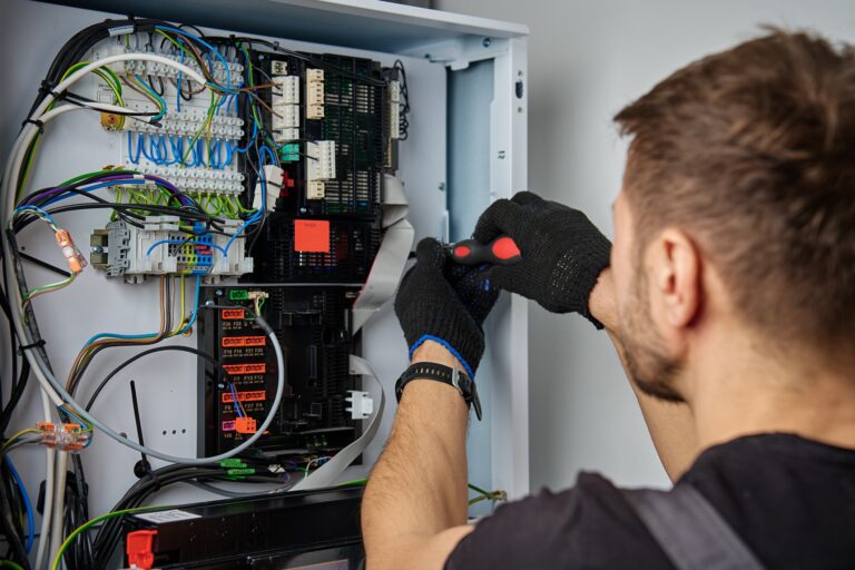 Eine Person mit Handschuhen arbeitet mit einem Schraubendreher an der Verdrahtung in einer offenen elektrischen Schalttafel.