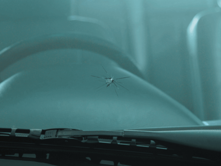 A close-up of a cracked windscreen with visible spiderweb-like damage, photographed from inside a car near the steering wheel and dashboard.