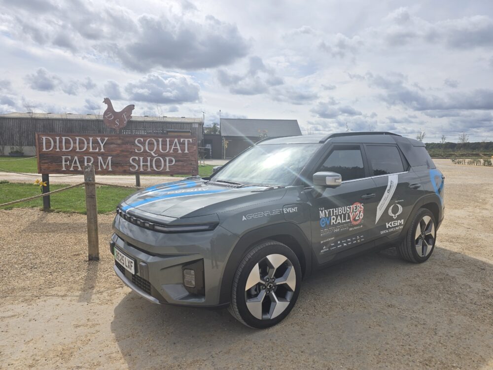 A grey SUV with “Mythbusters EV Rally 2026” decals is parked outside the Diddly Squat Farm Shop on a cloudy day, showcasing its participation in the exciting EV Rally.