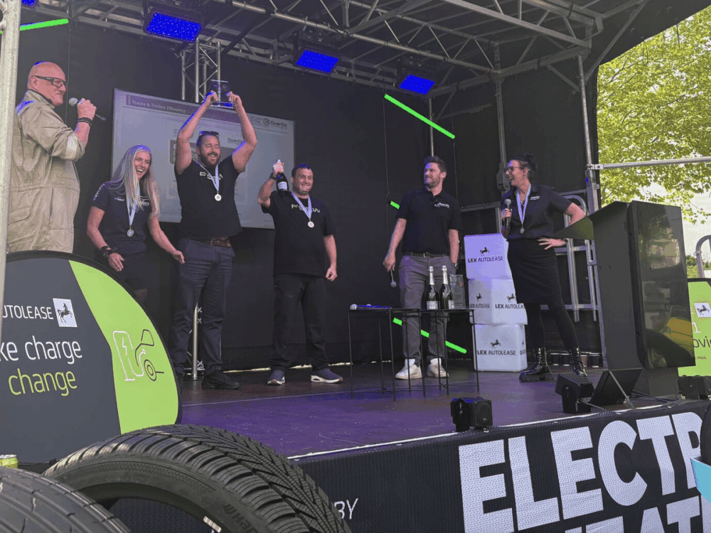 Five people on an outdoor stage celebrate with medals and a trophy at the EV Rally whilst an announcer speaks. Event branding and a tyre are visible in the foreground.