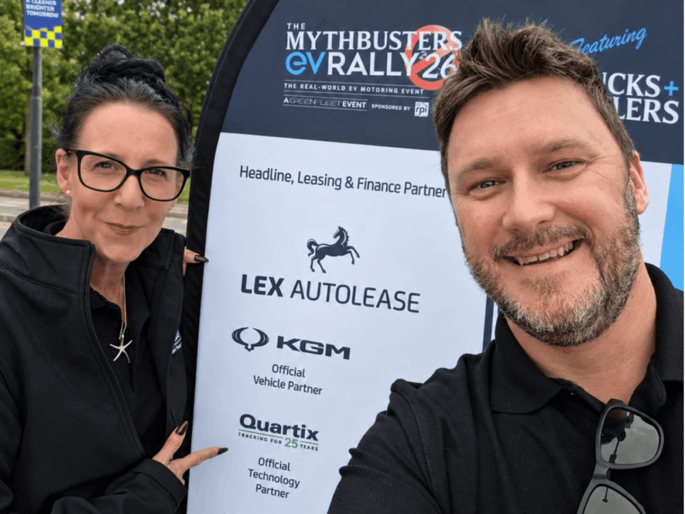 Two people stand next to a MythBusters EV Rally event sign displaying sponsors Lex Autolease, KGM, and Quartix.