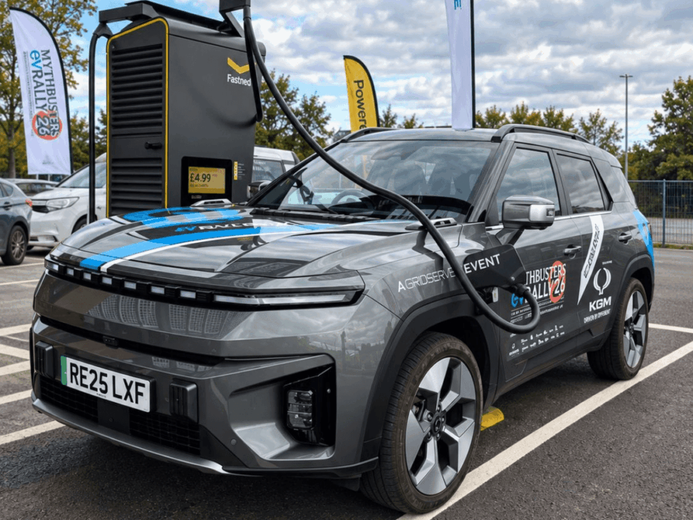 A grey electric SUV is parked at a charging point, plugged in and charging. The car features distinctive EV Rally event branding and rally stickers, with a visible number plate reading “RE25 LXF.”.
