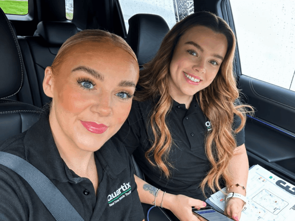 Two women wearing black Quartix polo shirts sit in a car during the EV Rally. One holds a mobile and a printed map, while both smile at the camera.