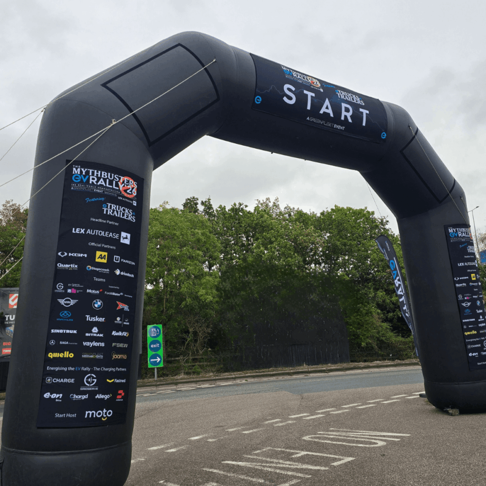 A large inflatable arch labelled “START” spans a road, proudly displaying sponsor logos on the sides for the EV Rally; trees and an overcast sky serve as the backdrop.