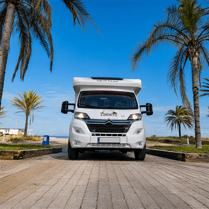 A white camper van is parked on a paved path lined with palm trees under a clear blue sky near a beach, showcasing the freedom and convenience that GPS de flotas para pymes españolas can offer for efficient travel and fleet management.