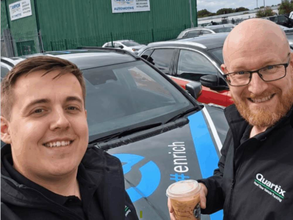Two people wearing Quartix jackets smile for a selfie in front of a car with blue markings at the EV Rally, one holding an iced coffee, in a car park with other vehicles and a green building in the background.