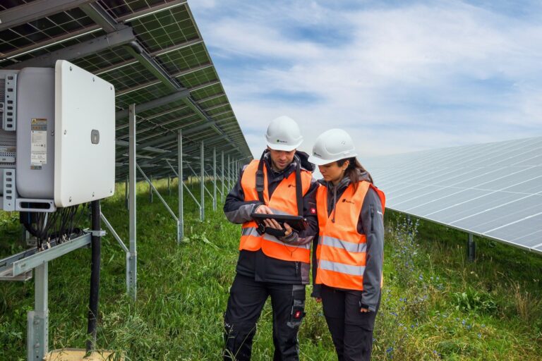 Dos técnicos, equipados con cascos y chalecos de seguridad, utilizan una tableta para la gestión de flotas de construcción mientras inspeccionan un parque solar.