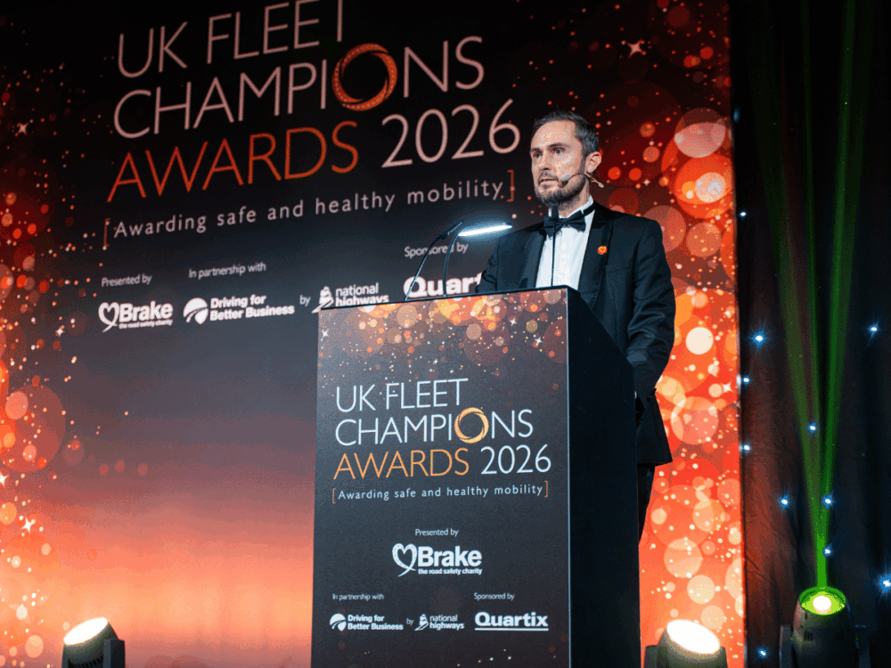 A man in a dinner jacket stands at a lectern on a stage with a backdrop for the UK Fleet Champions Awards 2026.
