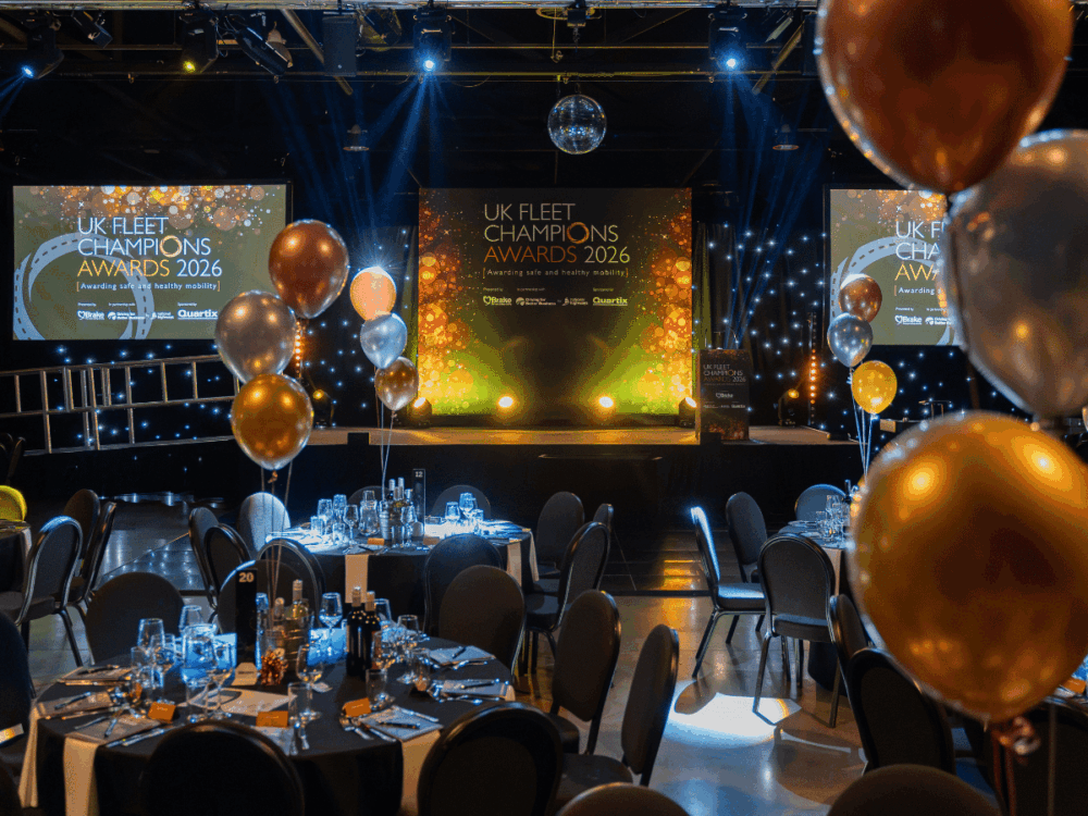 A banqueting hall set for the UK Fleet Champions Awards 2026, featuring a stage with screens displaying the event title and round tables decorated with gold and silver balloons.